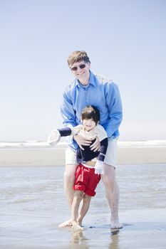 Father walking disabled son with cerebral palsy in the water at the ocean along beach