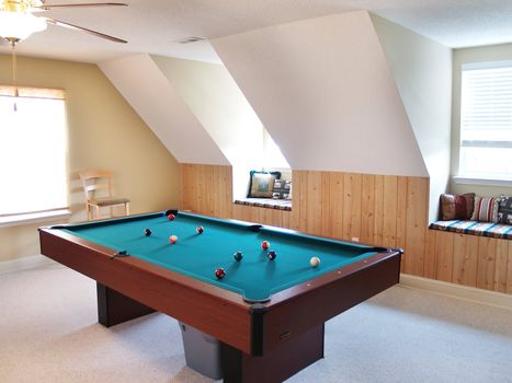 A pool table in a bonus room in a modern American juxury home. Two window seats in dormers are on the wall, and the balls are on the table waiting for a game.
