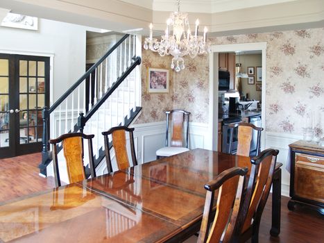 A long wooden table in a modern American luxury home. Six dining room chairs surround the table, and the stairs and foyer are visible in the background.