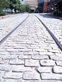 Train tracks embedded in down the middle of an old cobblestone road. The tracks curve into the distance in this perspective view.