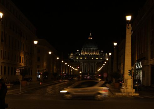 Traffic going by, with St. Peter's Basilica in the background, in Vatican city at night.
