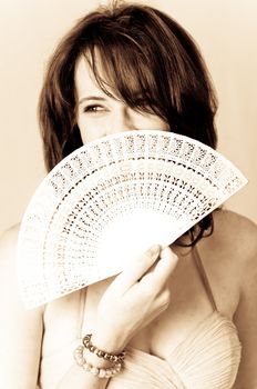 Sepia image of beautiful young brunette sexy model holding a white with Chinese hand fan in front of her face