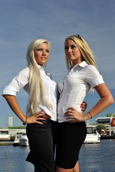 Two girls blonde stand on the pier. Boats on the background.