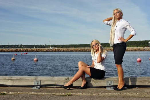 Two blonde girls on the beach. One is standing. Another sits