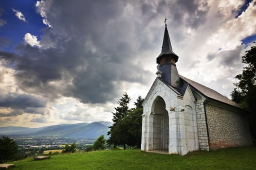 Wide-angle image of a little chapel, on a hillside in France, under wild cloudy skies. The sun behind the chapel.  Mountains in the distance with sun rays spreading over them. Space for copy in the sky.