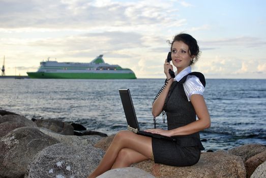 office by the sea: the girl on the rocks with a laptop and a desktop phone. ship on the backdrop