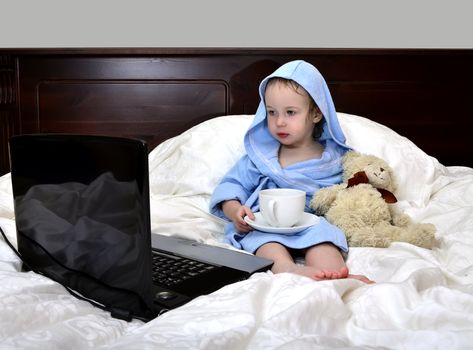 little girl in a bathrobe relaxing on the bed after a shower with cup of tea and laptop