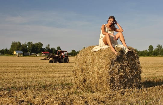 girl in a rural clothing sitting on the haystack, tractor on the background. space for text