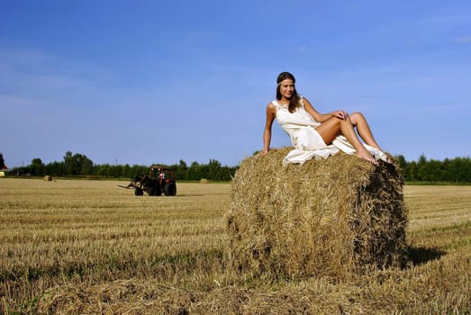 girl in a rural clothing sitting on the haystack, tractor on the background. space for text