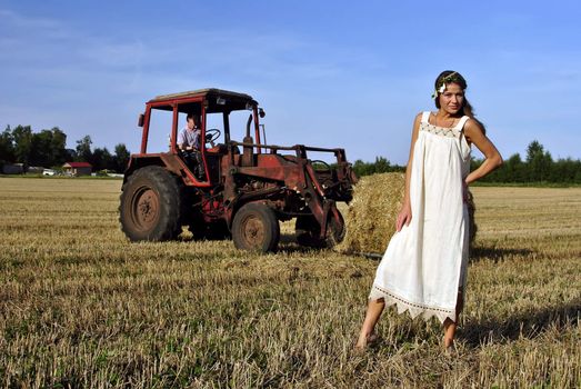 girl in a rural clothing standing on the field, tractor on the background