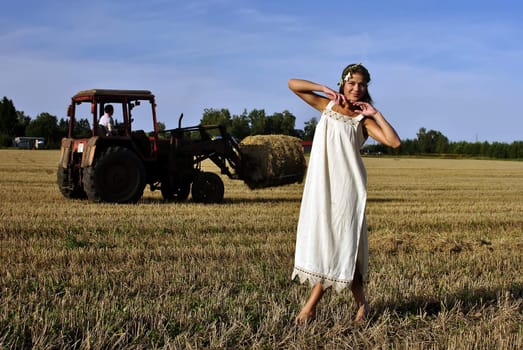 girl in a rural clothing standing on the field, tractor on the background