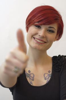 Portrait of a girl with red hair, piercing on lower lip, and tattoos on chest and shoulders, ok sign with hand.