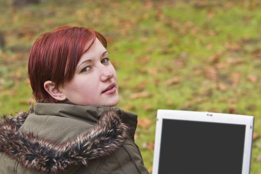 Redhead girl with computer in an autumn park looking up-back over the shoulder.