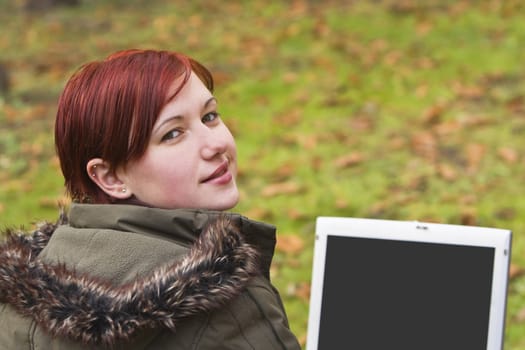 Smiling redhead girl with computer in an autumn park looking up-back over the shoulder.