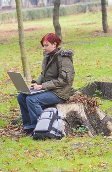 Redheaded girl working on computer in a park in autumn.