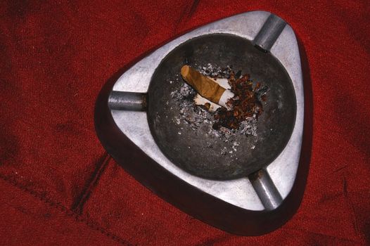 dirty metal ash tray with single stubbing sigaret in it on a red cloth
