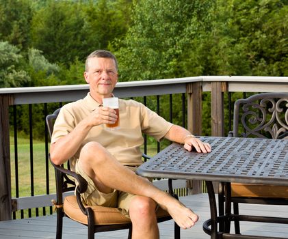 Middle aged man sitting on cast aluminium garden table on deck and drinking a glass of beer in back yard