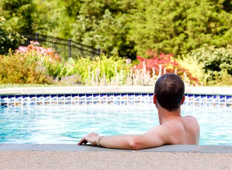 Senior male relaxing by the side of a modern swimming pool in back yard garden and facing away from the camera