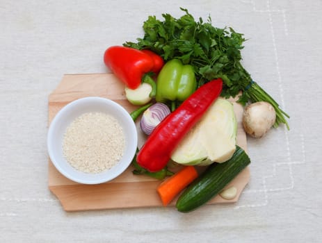 Upper view of various vegetables and a bowl of rice on a chopping board on a table.
