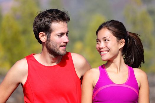Happy sporty couple portrait. Runners outside resting after running. Man and woman smiling at each other. Asian female athlete and white caucasian male model.