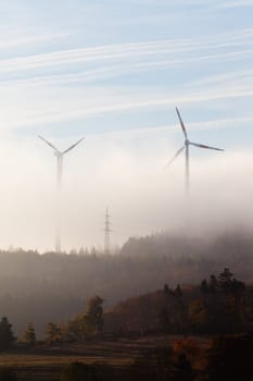 Ghostly shapes of large wind turbines dwarfing pylon of high-voltage transmission line on foggy morning in Eifel, rural Germany, Europe.