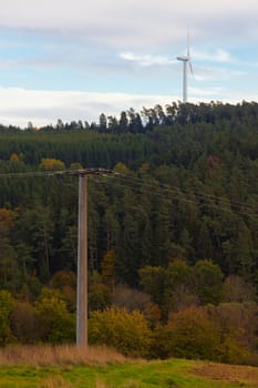 Large wind turbine on the horizon rising high above forest tree tops with high-voltage transmission line in foreground.