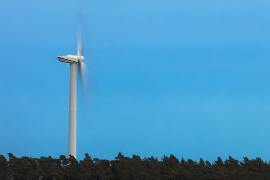 Green energy from wind: large wind turbine rotates fast (motion blur) high above forest tree tops.