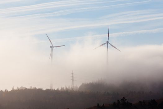Ghostly shapes of large wind turbines dwarfing pylon of high-voltage transmission line on foggy morning in Eifel, rural Germany, Europe.