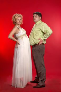 Kind of joke: who's belly is larger - pregnant woman's or her husband's? 
Young couple on red background.