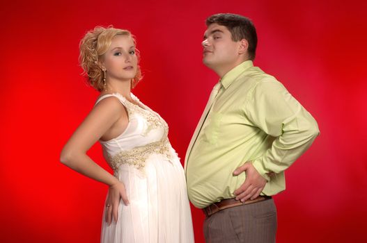 Kind of joke: who's belly is larger - pregnant woman's or her husband's? 
Young couple on red background. Horizontal version.