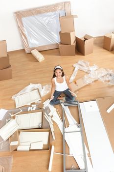 Moving woman in new home sitting on floor unpacking and assembling furniture.  Multiracial Asian Caucasian young woman.