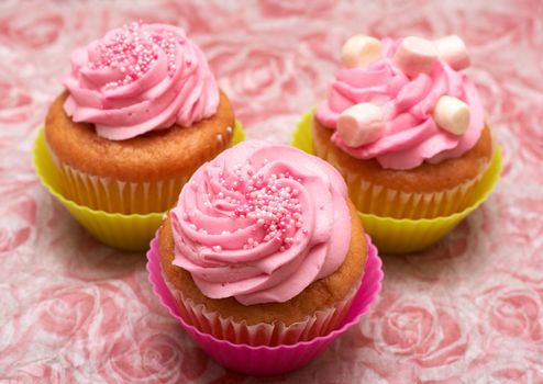 Three Fresh vanilla cupcakes in pink and green cups with strawberry icing, marshmallows and sprinkles on decorative background