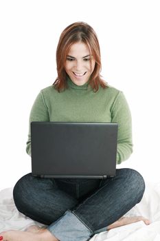 Excited girl in jeans with laptop sitting on her bed isolated