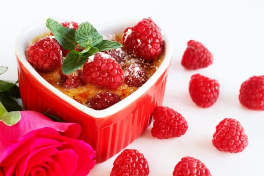 French creme brulee dessert with raspberries and mint covered with caramelized sugar in red heart shaped ramekin on white background