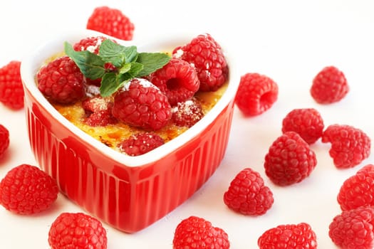 French creme brulee dessert with raspberries and mint covered with caramelized sugar in red heart shaped ramekin on white background