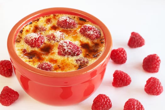 French creme brulee dessert with raspberries covered with caramelized sugar in red terracotta ramekin on white background