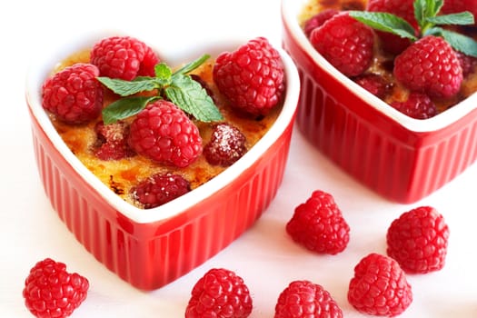 French creme brulee dessert with raspberries and mint covered with caramelized sugar in red  heart-shaped ramekins on white background