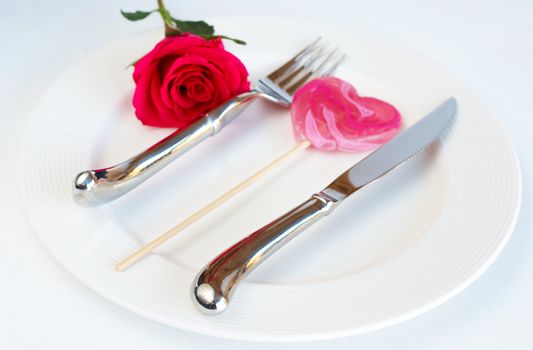 Valentines day table setting with a white plate, single pink rose and heart shaped lolly