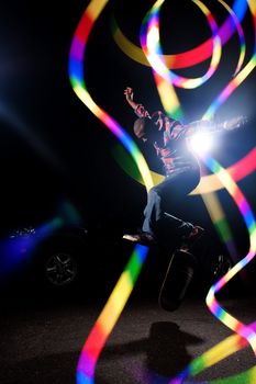 A young skateboarder doing jumping and kick flip tricks under dramatic rim lighting with lens flare and colorful rainbow light trails. 