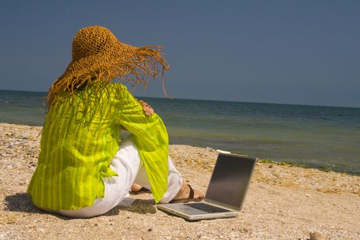 Woman in hat sitting on beach working on laptop