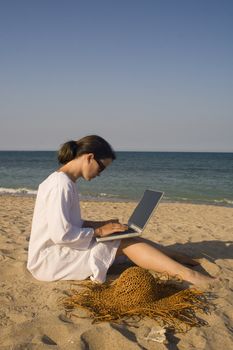 Woman sitting on beach in white dress working on laptop