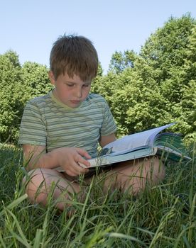 A little boy reads a big book with grass at background