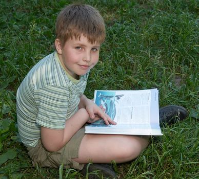A little boy reads a big book with grass at background
