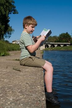 A little boy reads a big book with river at background