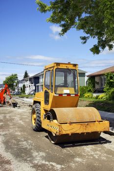 Construction, making pavement on the street of a small town and community neighborood: used yellow asphalt roller on dirt and gravel at the job site with mechanical digger and concrete sidewalk making in the background.