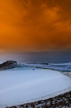 a snow covered links golf hole in ireland with blue flag and orange sunset sky