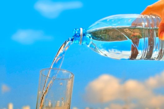 Water pours from a transparent bottle into a glass against a blue sky