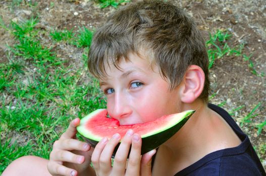 Boy eating watermelon slice holds it with both hands and looks askance at photographer