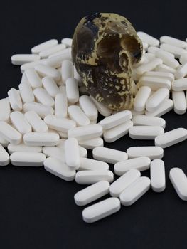 A small skull sits on a heap of white pills. Studio isolated over a black background.