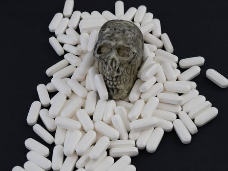 A small skull sits on a heap of white pills.  Studio isolated on a black background.